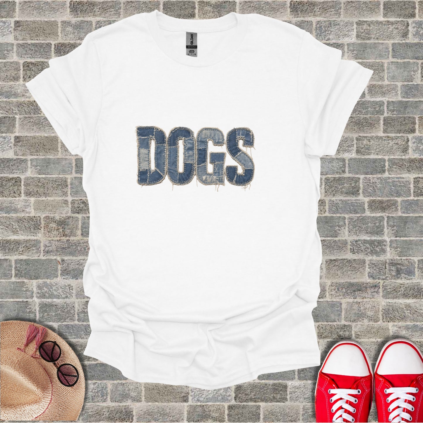 White t-shirt with "DOGS" text in denim-texture, laid flat with hat and sneakers.