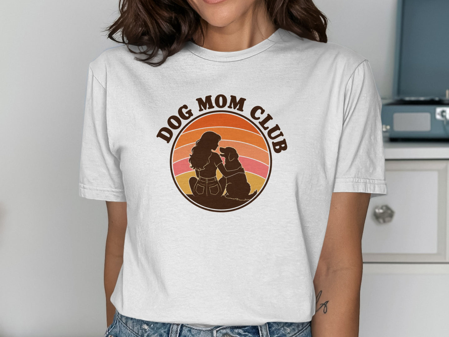 White t-shirt graphic with a silhouette of a woman and dog within a retro sunset circle that reads "DOG MOM CLUB."
