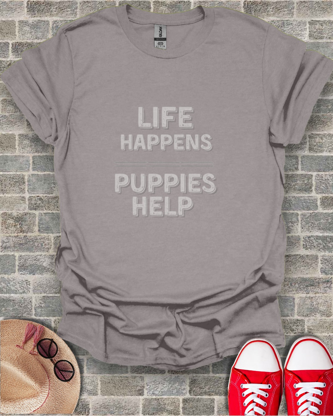 Gray t-shirt with text "LIFE HAPPENS PUPPIES HELP" against brick background.