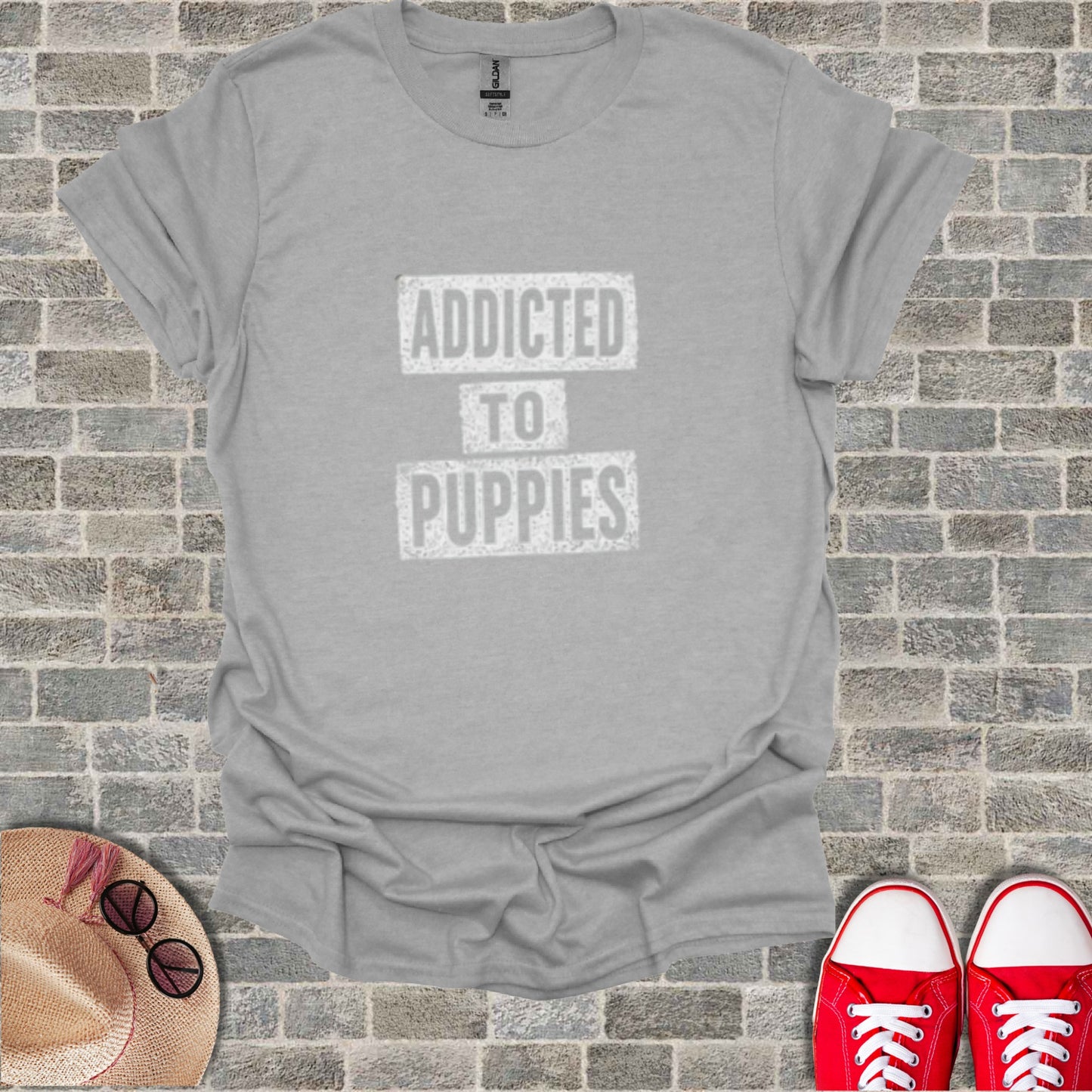 Gray t-shirt with "ADDICTED TO PUPPIES" text, flat lay with shoes and hat.