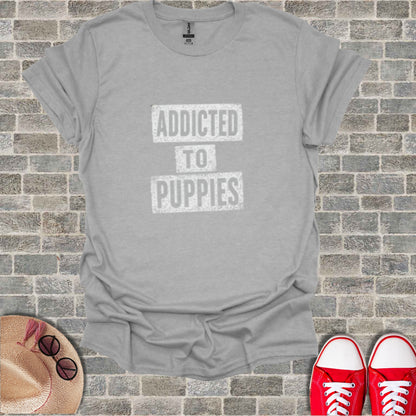 Gray t-shirt with "ADDICTED TO PUPPIES" text, flat lay with shoes and hat.