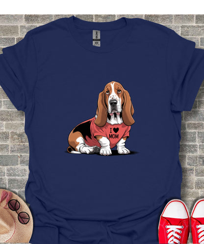 Basset hound wearing "I love Mom" t-shirt, sitting against brick wall.
