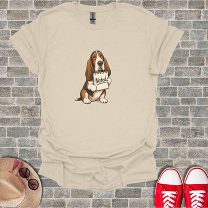 Basset hound sitting with a sign that reads "Noted."