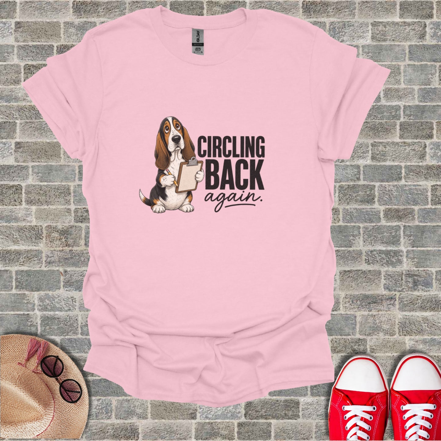 Pink t-shirt with a cartoon basset hound holding a clipboard that says "CIRCLING back again."