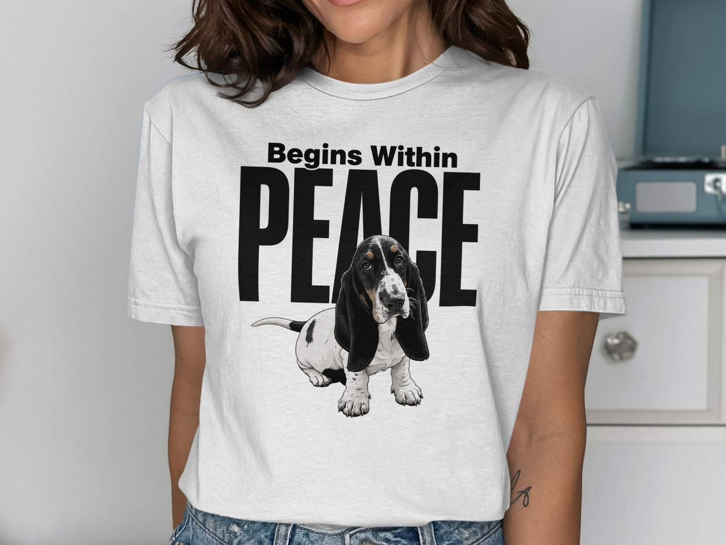 Basset Hound puppy sitting with "Begins Within PEACE" text on a white t-shirt.
