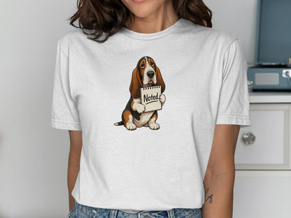 Basset hound wearing a white t-shirt holding a sign that says "Noted".
