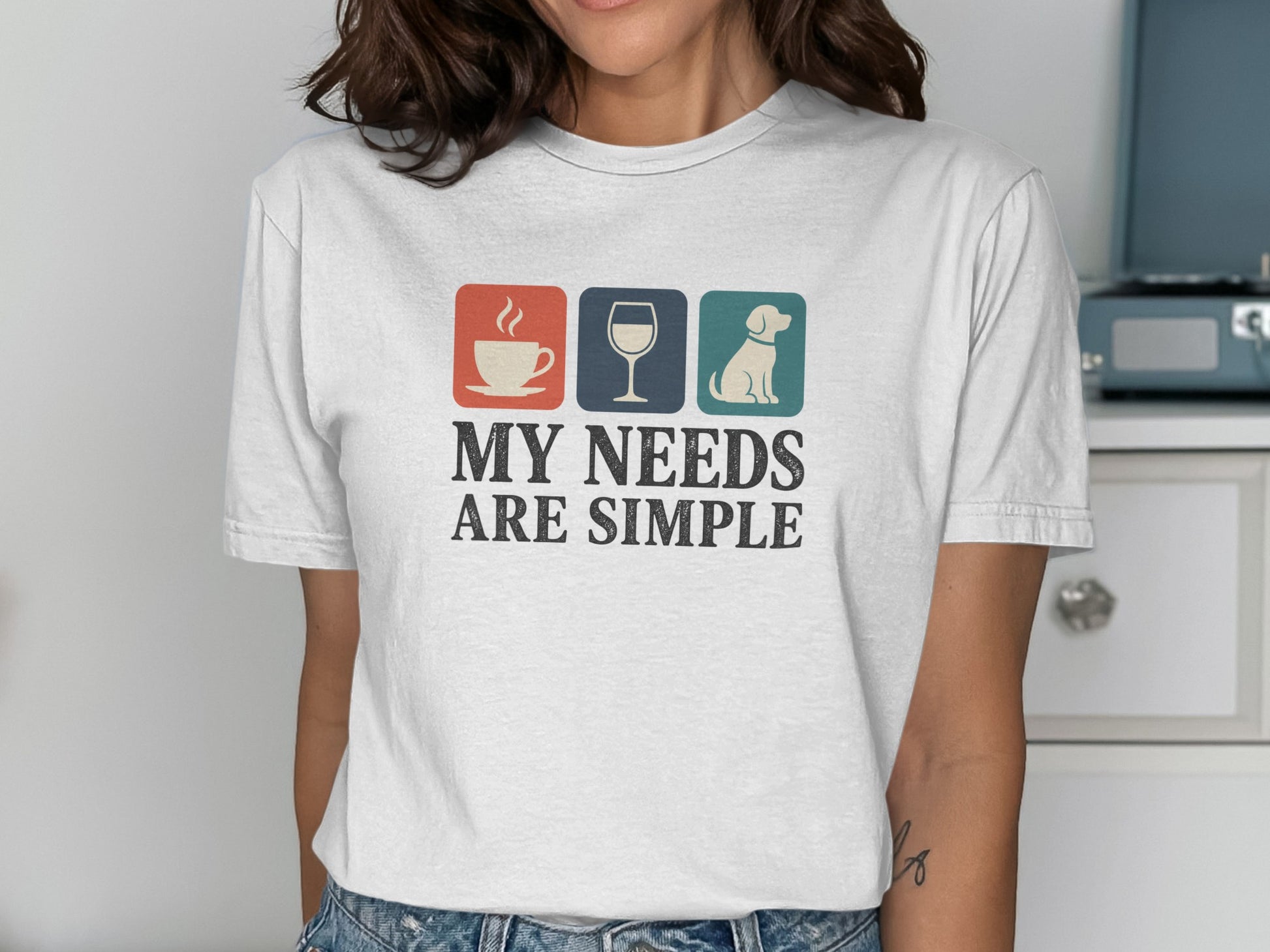 White t-shirt featuring icons of coffee, wine, and a dog with text "MY NEEDS ARE SIMPLE".
