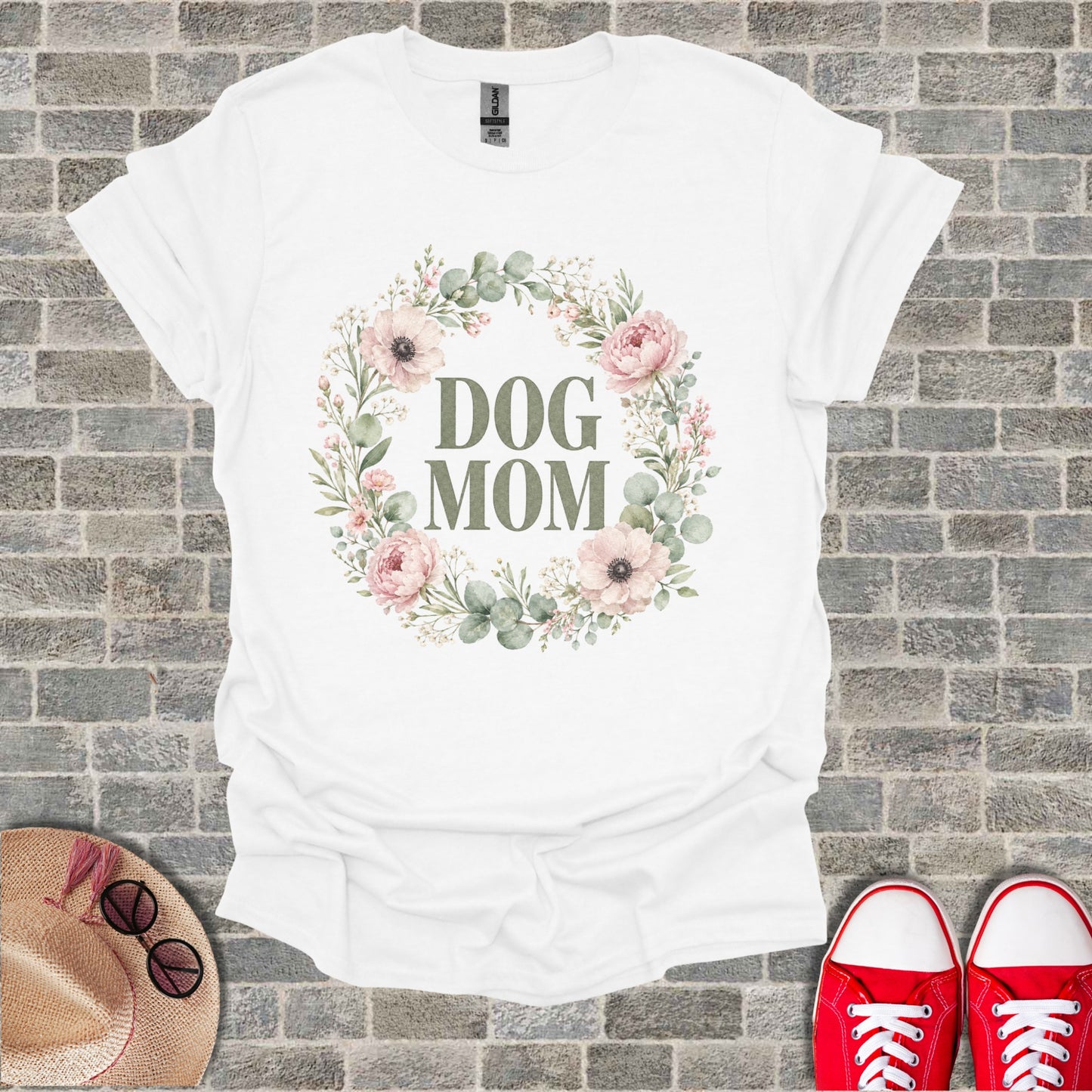 White t-shirt with "DOG MOM" text inside a floral wreath on a brick background.