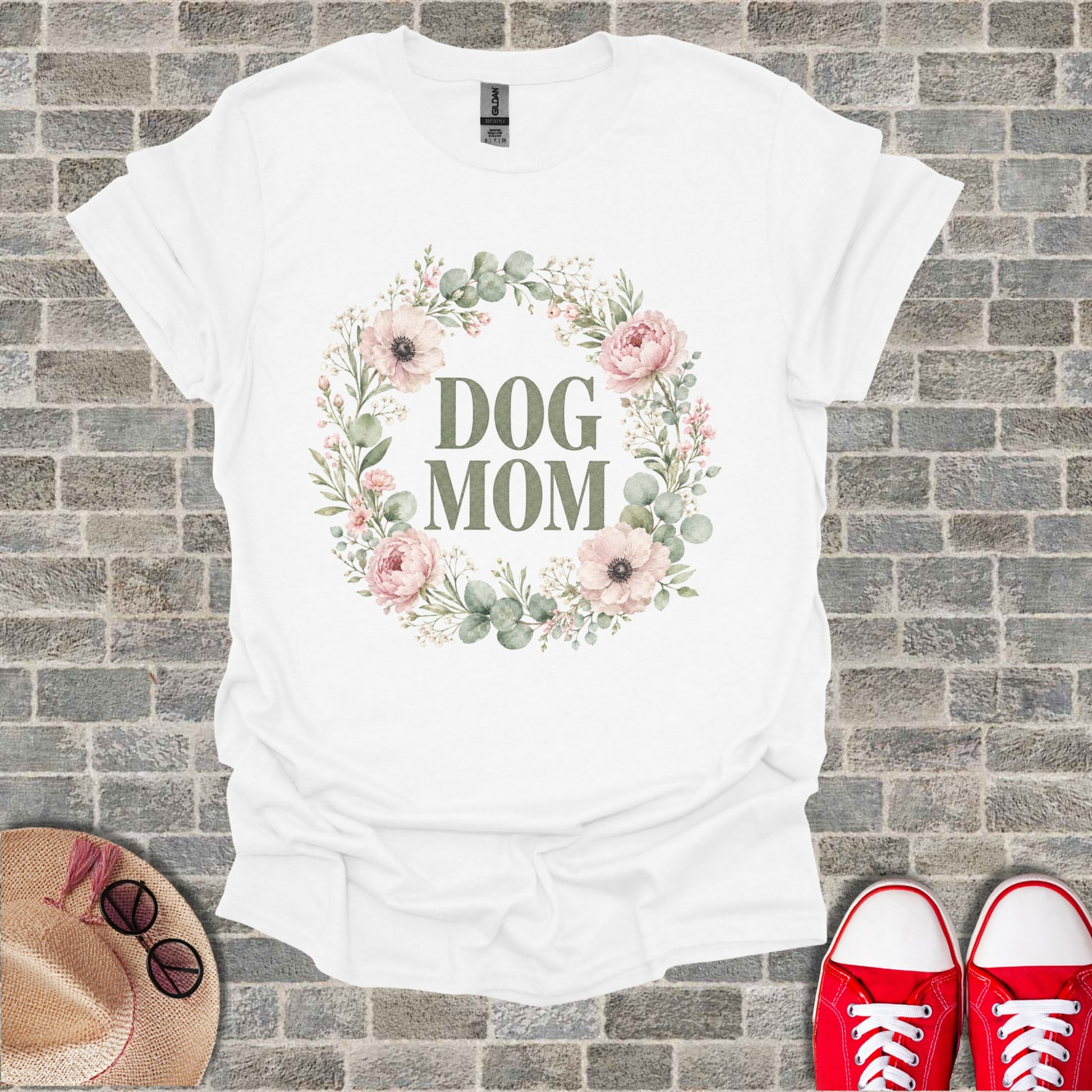 White t-shirt with "DOG MOM" text inside a floral wreath on a brick background.
