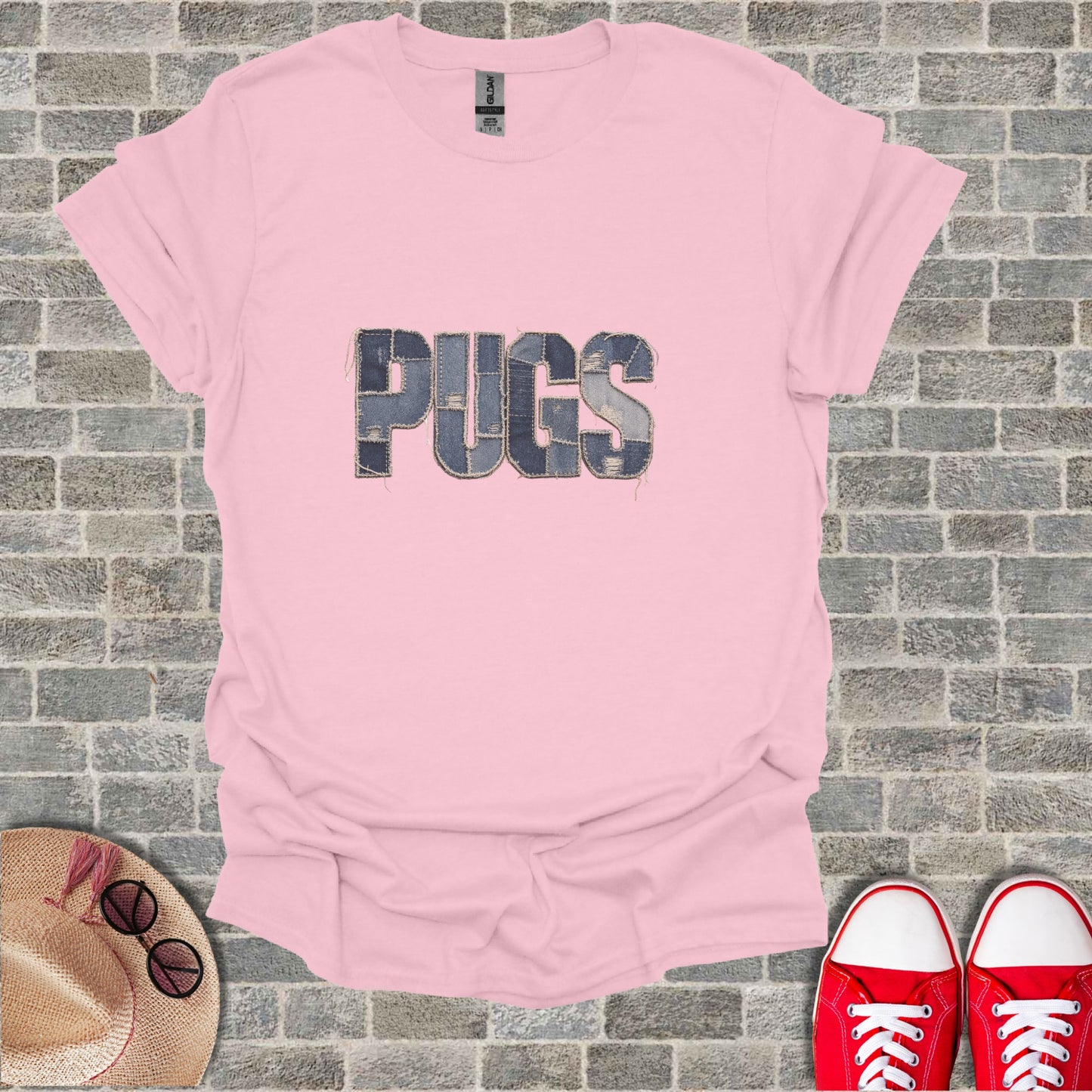 Pink t-shirt with "PUGS" text in denim patches and brick background.