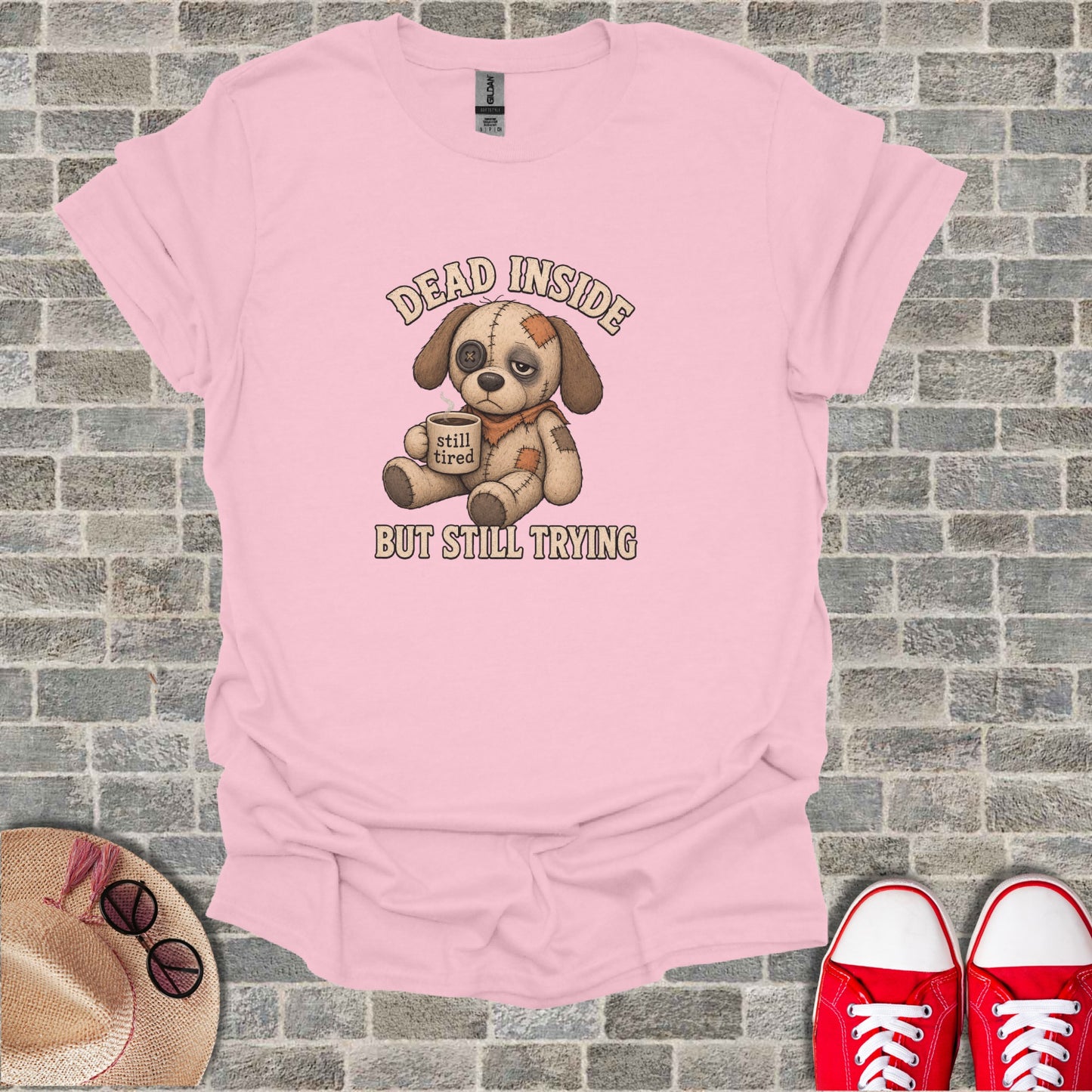 Pink t-shirt with a drawing of a sad dog holding a coffee cup and the text "DEAD INSIDE BUT STILL TRYING".