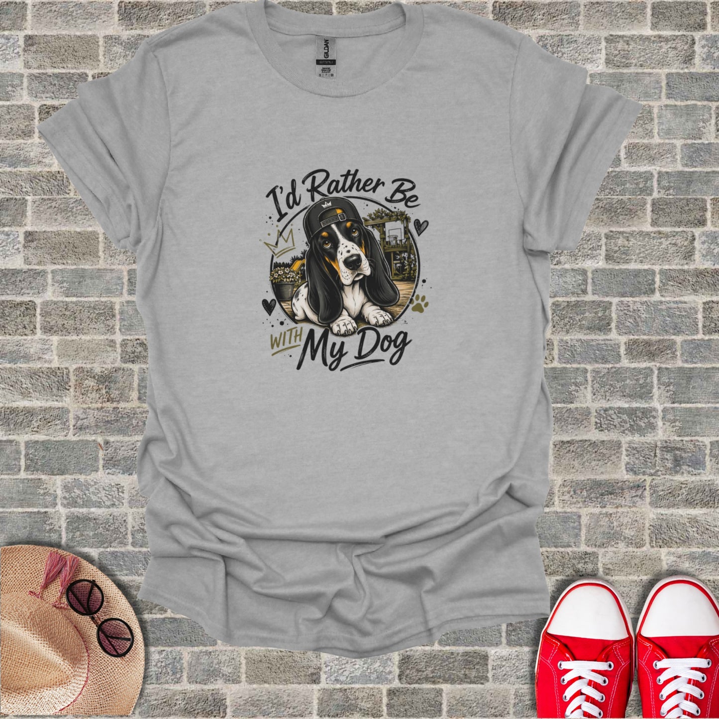 Basset Hound wearing a baseball cap and sunglasses, resting on a shirt that reads "I'd Rather Be With My Dog".