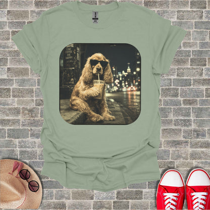Dog wearing sunglasses drinks from a cup on the street with city lights behind.