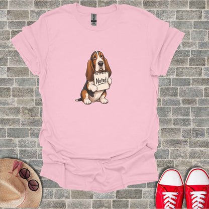 Pink t-shirt with a cartoon basset hound holding a "Noted." sign.